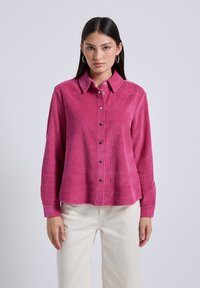 Camicia in corduroy rosa con bottone frontale, maniche lunghe e bottoni neri. Presenta un colletto strutturato e una texture a costine.