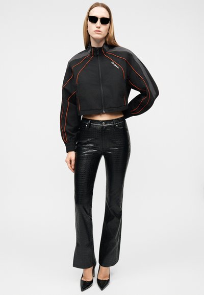 Γυναίκα που φοράει μαύρο κούμπωμα με φερμουάρ στο ύψος του κοψίματος με πορτοκαλί ρίγες, μαύρο υφασμάτινο παντελόνι με ψηλή μέση, μαύρες μυτερές γόβες και μαύρα γυαλιά ηλίου.