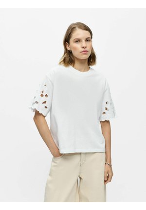 Kobieta o krótkich włosach, ubrana w biały top z krótkim rękawem z wycięciami w kształcie kwiatów oraz jasnobeżowe spodnie, z ręką w kieszeni, neutralne tło.