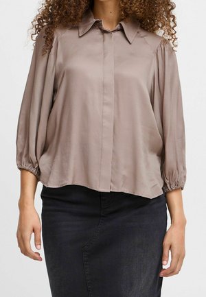 Žena v volné, světle hnědé blůze na knoflíky s nafouknutými tříčtvrtečními rukávy a límečkem, v kombinaci s tmavou sukní.