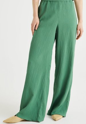 Femme portant un pantalon large vert texturé et des ballerines pointues beige tressées, debout devant un fond uni.