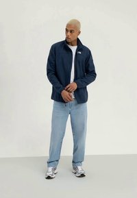 Casaco de lã azul-marinho com fecho, t-shirt branca, jeans azul-claro e ténis prateados. Modelo em pé com os braços cruzados, fundo neutro.