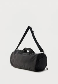 Μαύρη σακούλα duffel κατασκευασμένη από ανθεκτικό ύφασμα, με διπλές λαβές μεταφοράς, ρυθμιζόμενο ιμάντα ώμου και φερμουάρ σε πλαϊνή θήκη.