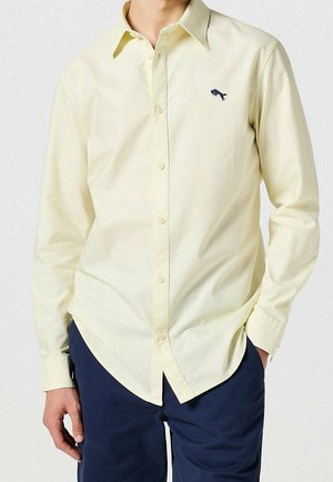 Homme portant une chemise jaune pâle à manches longues avec un petit logo brodé foncé sur la poitrine et un pantalon bleu marine.