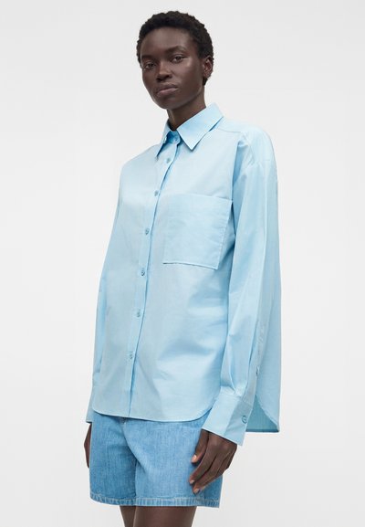 Γυναίκα που φορά υπερμεγέθη ανοιχτομπλε πουκάμισο με τσέπη στο στήθος και ανοιχτομπλε τζιν σορτς σε απλό φόντο.