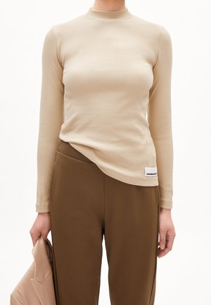 Nainen, joka pukeutunut beigeen, ribbineulottuun pitkähihaiseen paitaan ja ruskeisiin korkeavyötäröisiin housuihin, vasemmassa kädessään beigeä bleiseriä.