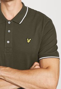 Olivově zelená polo košile vyrobená z texturované látky. Má kontrastní bílý límec a lemy rukávů. Žluté vyšívané logo na prsou.