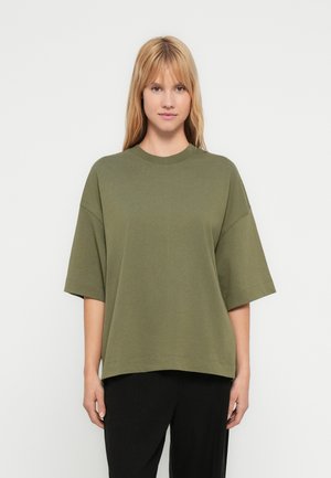 Maglietta di cotone verde oliva con vestibilità rilassata, maniche corte e scollo rotondo, abbinata a pantaloni neri larghi.