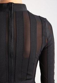 Musta paita, jossa on pystysuoria raidoitettuja läpinäkyviä ja kiinteitä paneeleja, korkea kaulus ja takana vetoketju. Tasainen pinta yhdistettynä jämäköihin muotoiluelementteihin.