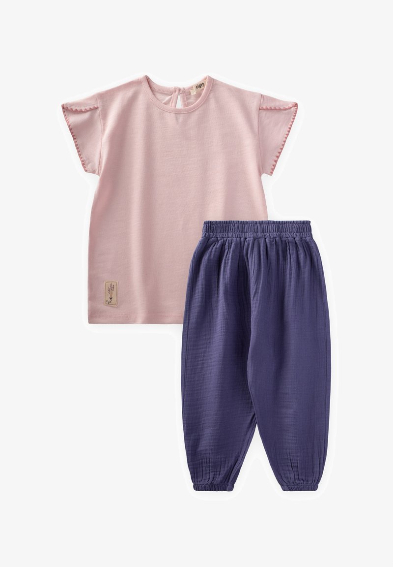 Šviesiai rožinė medvilninė viršutinė dalis su trumpomis rankovėmis ir banguotais kraštais, derinama su tamsiai violetinėmis tekstūruotomis kelnėmis su elastinga juosmena. Ant viršaus matomas etiketė.