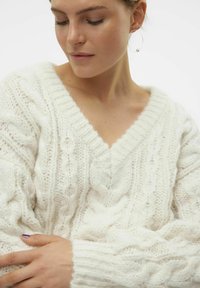 Pull en maille côtelée couleur crème avec un col en V et un fil épais et texturé. Le design présente des motifs en relief et des poignets côtelés.