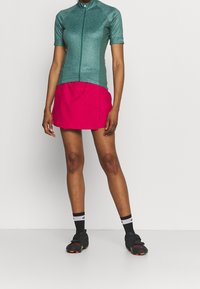 Kortärmad cykeltröja i teal med ett vågmönster, matchad med en ljusrosa skorts. Inkluderar svarta sportskor och randiga strumpor.