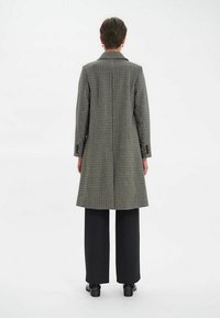 Manteau en laine à carreaux avec une silhouette longue et des revers crantés, présentant une fente dans le dos, associé à un pantalon noir à jambes larges.