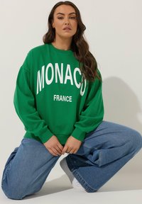 Zielona bluza z białym napisem "MONACO FRANCE", o luźnym fasonie i długich rękawach. Noszona z luźnymi niebieskimi dżinsami i białymi sneakersami.