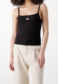 Top a righe nere con spalline sottili, caratterizzato da una piccola toppa con il logo di Tommy Jeans. Abbinato a pantaloni leggeri, texturizzati e plissettati.