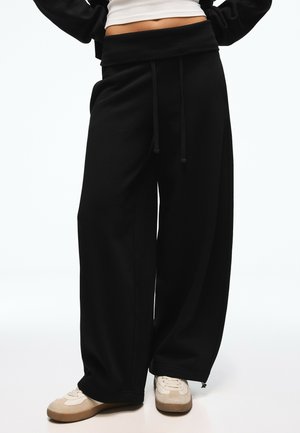 Personne portant un pantalon noir à jambes larges avec cordon à la taille et des baskets blanches, debout les mains sur les hanches.