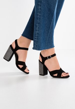 Czarne zamszowe sandały na wysokim obcasie z krzyżującymi się paskami i regulowanym zapięciem na kostce. Blokowy obcas ozdobiony jest rzędami srebrnych ćwieków.