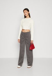 Maglione di maglia crema tagliato, pantaloni grigi a gamba larga e una borsa rossa. Modella che indossa sneakers bianche. Texture morbide e linee pulite.