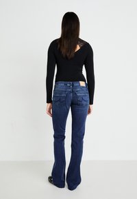 Dunkelblaue, ausgestellte Jeans mit hoher Taille, ausgestattet mit zwei Gesäßtaschen und einem Lederlogo-Patch am Bund. Schwarzes Langarmoberteil mit Spitzendetails.