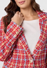 Rood, blauw en geel geblokte blazer met een notch-kraag en drie gouden knopen op de manchetten, gelaagd over een wit top.