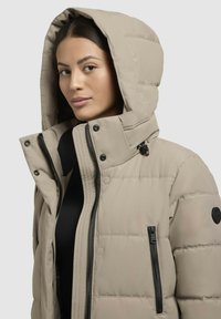 Beige pufferjakke med hætte, der har sorte lynlåse, en lynlåslomme og en struktureret overflade. Stylet med en høj krave.