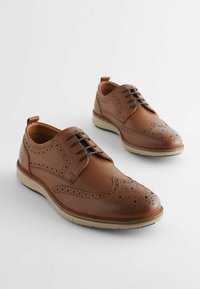 Chaussures habillées en cuir marron avec des perforations décoratives, bout rond, lacets noirs et semelle en caoutchouc de couleur claire. Texture lisse avec un design classique.