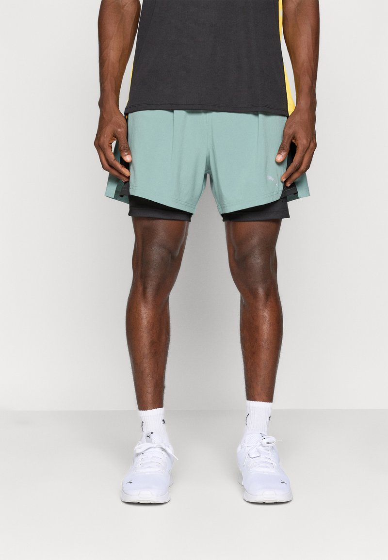 Pantalones cortos deportivos en verde claro superpuestos sobre pantalones cortos de compresión negros. Zapatillas blancas con sutiles acentos y calcetas blancas con patrón.