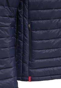 Section de veste matelassée bleu marine avec poche verticale zippée et petite étiquette de marque rouge en tissu près du bord inférieur.