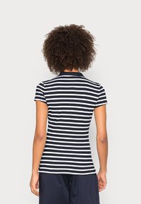 Personne aux cheveux bouclés portant un t-shirt à manches courtes à rayures horizontales noires et blanches et un short foncé, tourné(e) de dos à la caméra.
