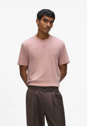 Homme portant un t-shirt à manches courtes rose pâle avec un petit logo et un pantalon plissé marron foncé, debout les mains derrière le dos.