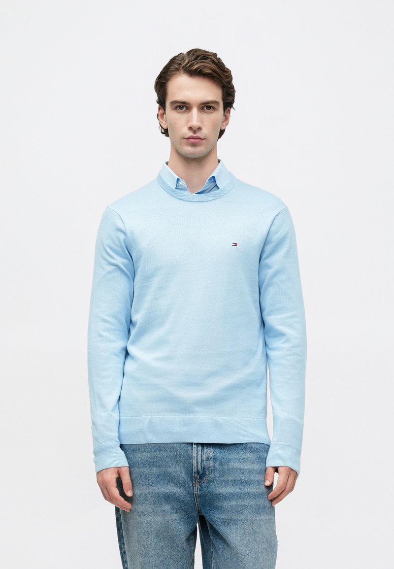 Egy fiatal férfi állva, szemben nézve, világoskék pulóvert visel egy világoskék galléros ing felett, valamint laza kék farmert, fehér háttér előtt.