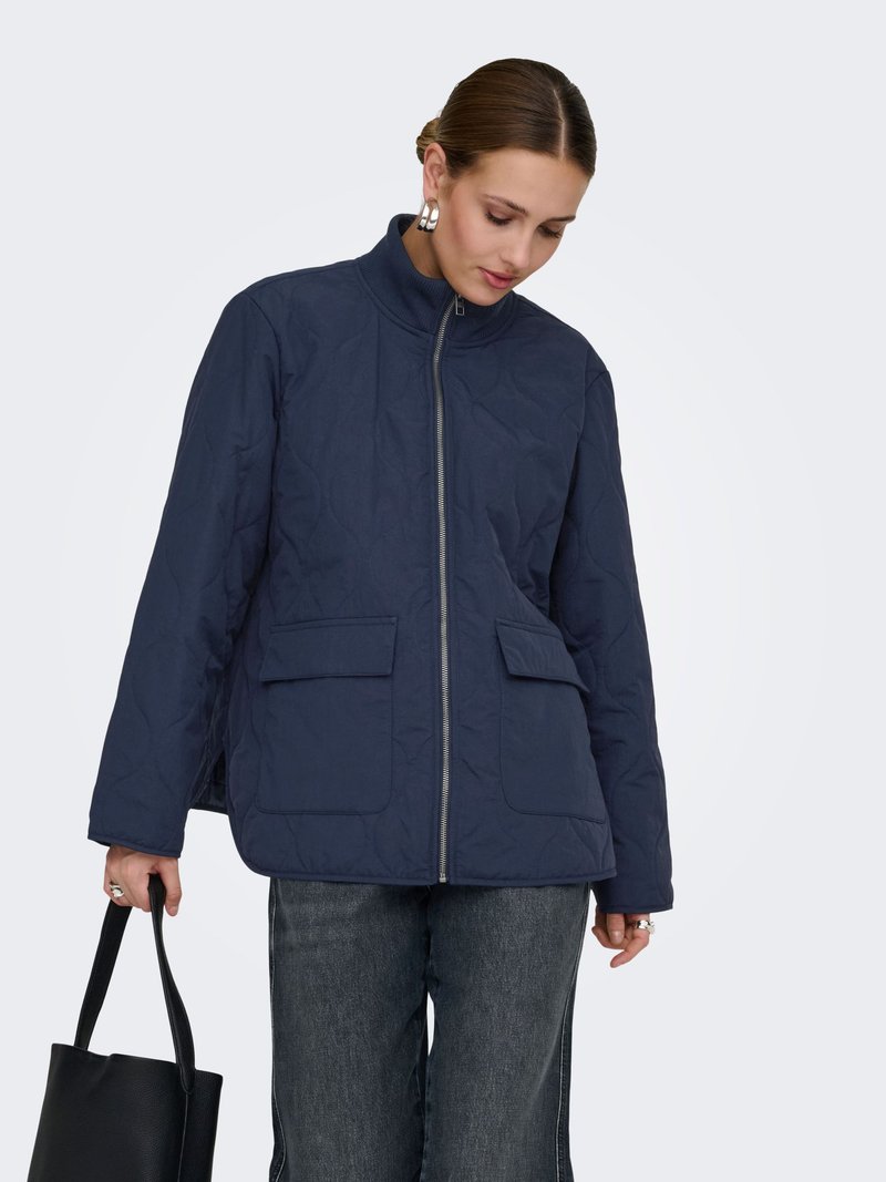 Femme portant une veste matelassée bleu marine avec poches avant à rabat, tenant un sac cabas noir et regardant vers le bas devant un fond uni.