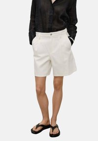 Shorts en lin blanc avec une coupe ajustée, des poches latérales et une fermeture à bouton, associés à une chemise noire à manches longues en voile et des tongs noires.