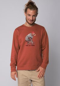 Sweat-shirt couleur rouille à manches longues présentant une illustration d'un ours en tenue rayée faisant du monocycle. Le texte en dessous indique "BOURREAU DE FARCE."