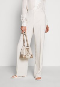 Pantalon taille haute beige à jambe droite, associé à un chemisier blanc. Tient un sac seau beige et crème avec des accents dorés. Haut à manches longues avec des poignets.