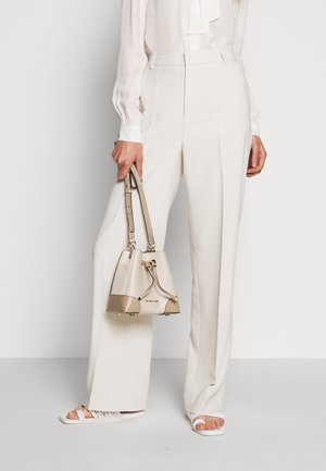 Pantaloni beige a vita alta con gamba dritta, abbinati a una blusa bianca. Tiene in mano una borsa a secchiello beige e crema con dettagli dorati. Maglia a maniche lunghe con polsini.