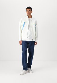 Balta zip-up striukė su mėlynomis ir žaliomis detalėmis, derinama su mėlynomis džinsinėmis kelnėmis ir baltomis sportinėmis bateliais. Medžiaga atrodo lygi ir lengva.