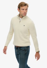 Ivo-white pullover met een hoge kraag, rits, ribbelmanchetten en een klein logo op de borst, gecombineerd met donkerblauwe jeans.