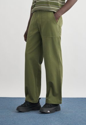 Personne portant un pantalon large couleur vert olive, des chaussures de randonnée noires, et une chemise rayée verte, debout sur un tapis bleu avec une main dans la poche.