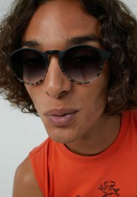 Gafas de sol redondas negras con lentes degradados, con un borde estampado. El modelo lleva una camiseta sin mangas naranja. Se ve el cabello rizado.