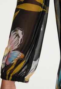 Tejido negro translúcido con patrones florales y abstractos coloridos. Manga larga abullonada con detalle de puño elástico. Ligero y texturizado.