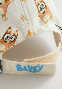 Casquette blanche ornée de personnages de dessin animé dans des tons d'orange et de crème, avec une texture douce. La sangle arrière est brodée de "Bluey" en bleu.