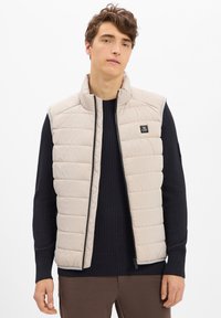 Junger Mann, der eine beigefarbene, gesteppten ärmellosen Weste über einem schwarzen Rippenpullover und braunen Hosen trägt, steht mit neutralem Ausdruck vor einem weißen Hintergrund.