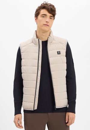 Jonge man in een beige gewatteerd mouwloos vest over een zwart geribbeld sweater en een paarse broek, staand met een neutrale uitdrukking tegen een witte achtergrond.