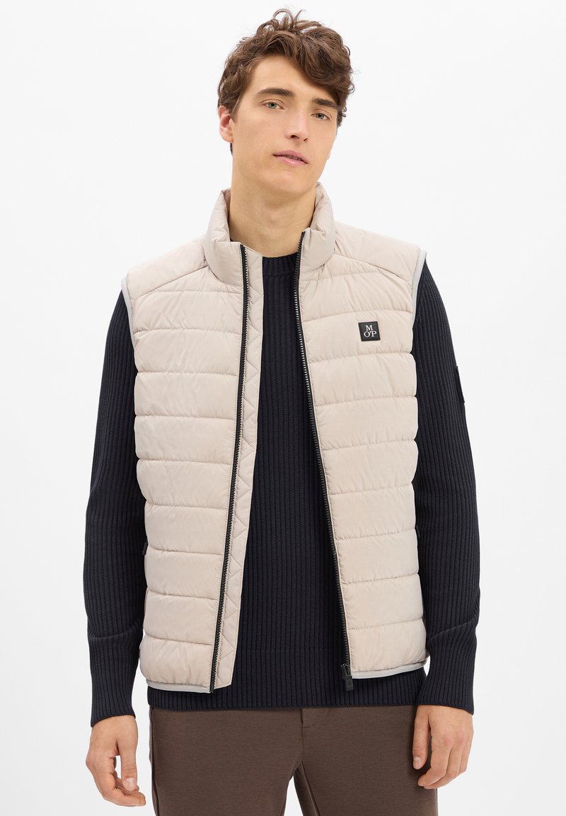 Junger Mann, der eine beigefarbene, gesteppten ärmellosen Weste über einem schwarzen Rippenpullover und braunen Hosen trägt, steht mit neutralem Ausdruck vor einem weißen Hintergrund.