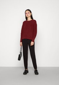 Burgundska rebrasta pulover s spuštenimi rameni, v kombinaciji z črnimi oprijetimi hlačami. Model drži majhno črno teksturirano torbico in nosi roza sončna očala.