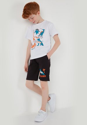 Ragazzo con capelli rossi che indossa una maglietta bianca con stampa di dinosauri e pantaloncini neri con motivo di dinosauri, in piedi contro un muro bianco.