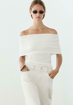 Frau trägt weißes schulterfreies Oberteil, helle Hose, schwarze Sonnenbrille, Hände in den Taschen vor einfarbigem Hintergrund.