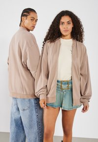 Two models wear oversized, beige sweatshirts. The male model has braided hair and denim pants, while the female model pairs her sweatshirt with a belted denim skirt.