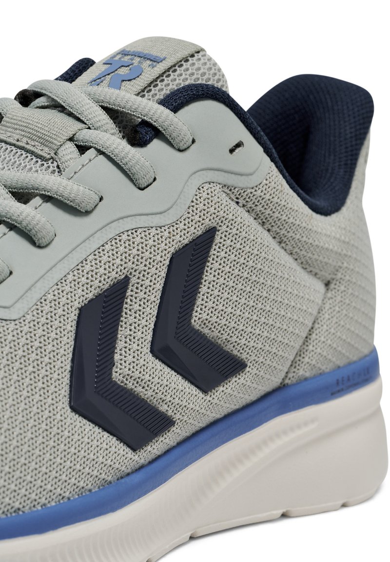 Zapatilla deportiva gris claro con una parte superior de malla texturizada, detalles en azul marino y patrones de chevron azul oscuro en el lateral. Suela acolchada con un ribete azul.